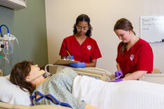 Two nursing students conducting tests on a mannequin in a hospital bed.